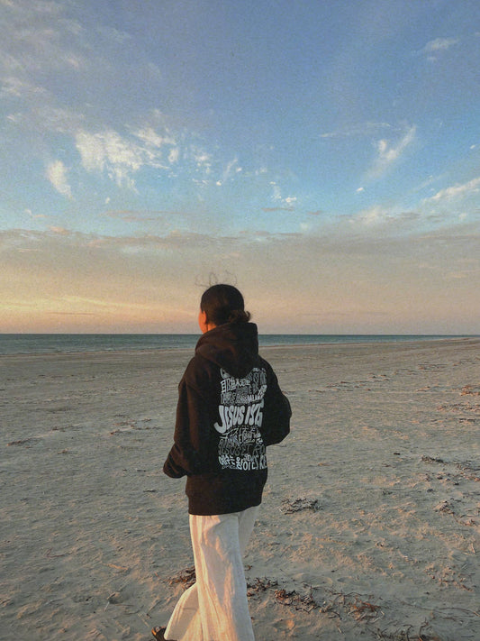 Woman wearing the Jesus Is King Hoodie at sunrise on the beach, showcasing the multilingual “Jesus Is King” back graphic while resting her hands in the front pouch pocket.