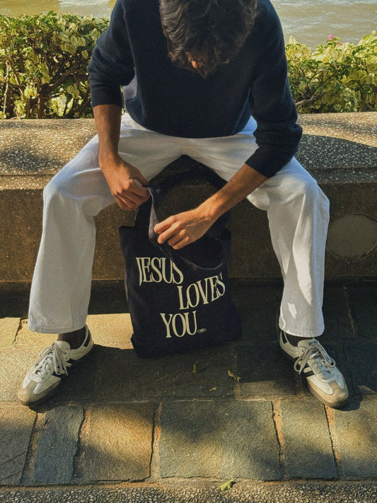 Person sitting on a stone bench holding a navy blue Christian tote bag with 'Jesus Loves You' text.