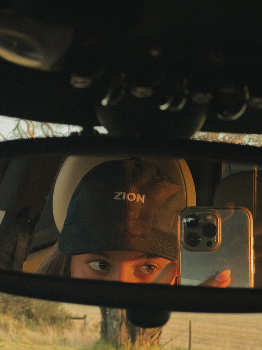 Rear-view mirror selfie of a woman wearing the dark green Zion Canvas Cap during golden hour, highlighting the textured canvas fabric and embroidered Zion logo.