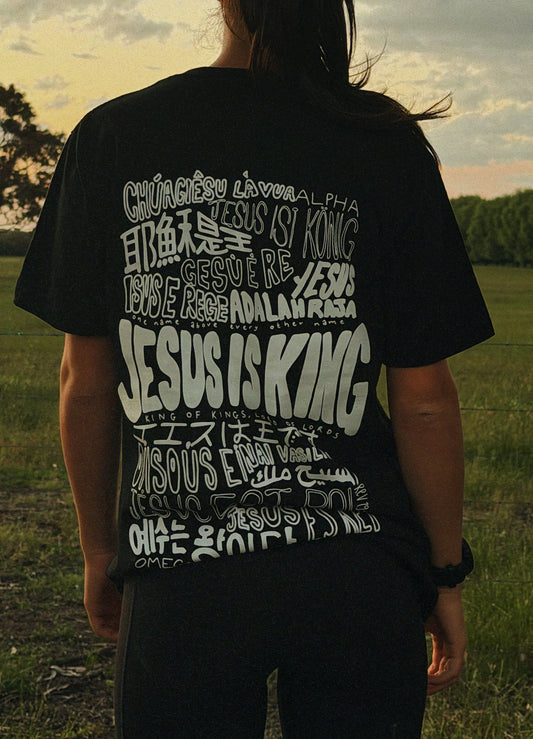 Woman wearing the Jesus Is King black Christian t-shirt by Zion Christian Clothing with the original multilingual back graphic displayed outdoors.