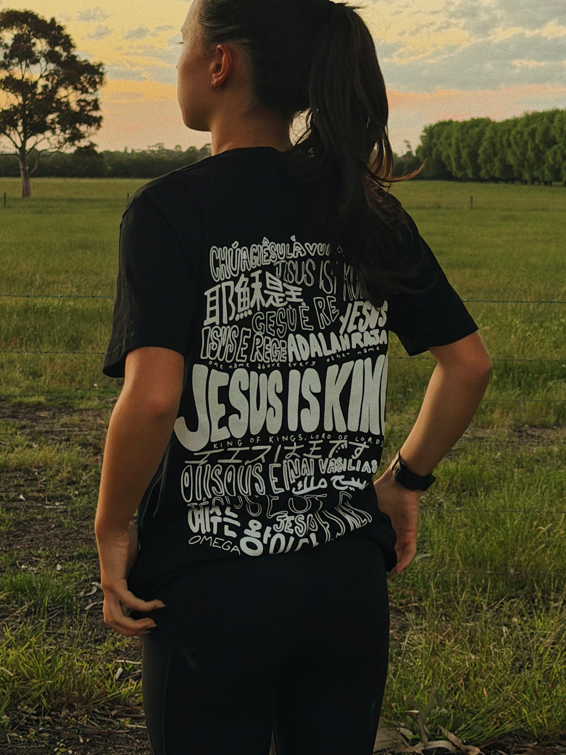 Lifestyle photo of a woman wearing the Jesus Is King black Christian t-shirt with the original multilingual back graphic while outdoors.